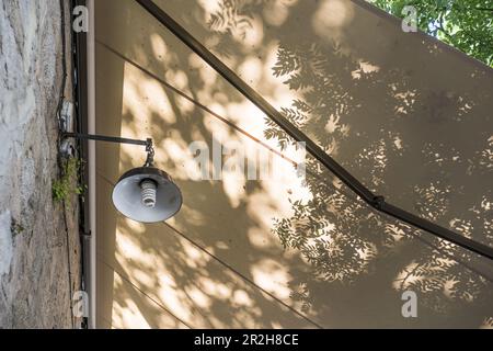 Eine Außenfassadenleuchte unter einer ausziehbaren Pergola und mit vielen schattigen Ästen Stockfoto