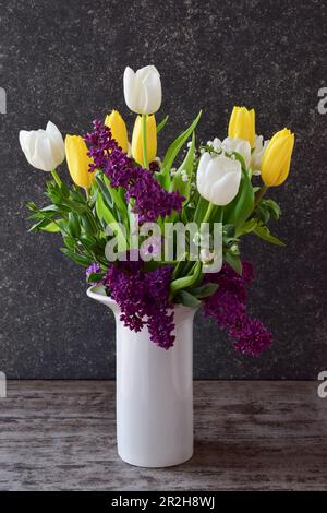 Ein wunderschöner Strauß aus Tulpen und Fliedern in einer weißen Vase vor dunklem Steinhintergrund, Seitenansicht Stockfoto