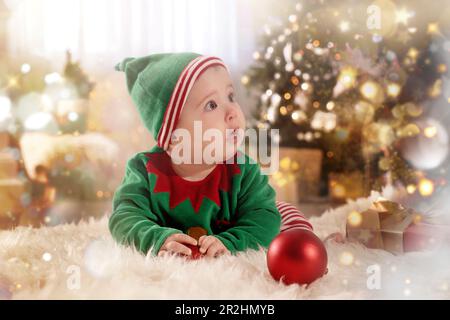 Baby trägt süßes Elfenkostüm auf dem Boden im Zimmer, das zu Weihnachten dekoriert ist. Magische festliche Atmosphäre Stockfoto