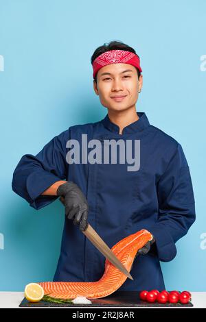 Lächelnder junger Mann in blauer Uniform, der im Studio Fischfilet kocht, ein Messer in einer Hand hält, leckeres Essenskonzept, Kopierraum Stockfoto