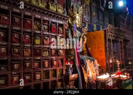 Buddha-Figur und antike Wandeinheit mit Kerzen und Opfern im dunklen Inneren eines Gebetssaals im Kumbum Champa Ling Kloster, Xining, China Stockfoto