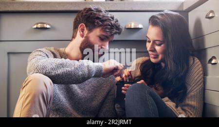 Eiscreme, entspannen Sie sich und ein Paar auf dem Boden in der Küche, um sich zu verbinden, zu entspannen und eine schöne Zeit zusammen zu verbringen. Liebe, Beziehung und Mann und Frau, die süß miteinander teilen Stockfoto