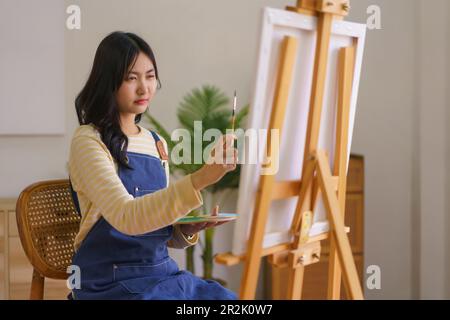 Junge Künstlerin, die Proportionen mit Pinsel auf Leinwand auf Staffelei im Studio misst. Stockfoto