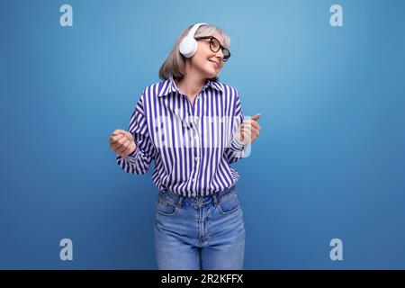 Energiegeladene Rentnerin mit grauen Haaren, die sich mit Musikgeräten auf hellem Studiohintergrund vertraut macht Stockfoto