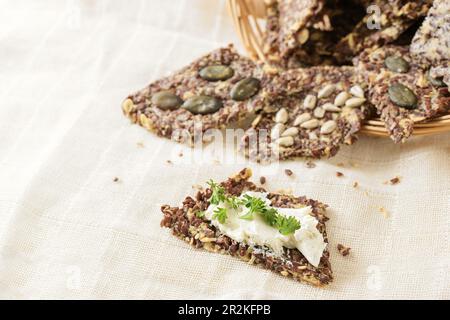 Gesunde Snacks aus Leinsamen, Hafer, Kräutern und verschiedenen Samen, einer mit Frischkäse und Petersilie auf einer hellen beigen Serviette, Kopierraum, ausgewählter Fokus Stockfoto