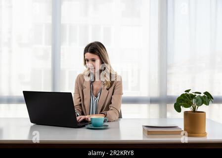 Junge, glückliche Geschäftsfrau, die am Schreibtisch sitzt und an einem Laptop in einer modernen Büroeinrichtung arbeitet. Lächelnde Mitarbeiterin mit CO Stockfoto