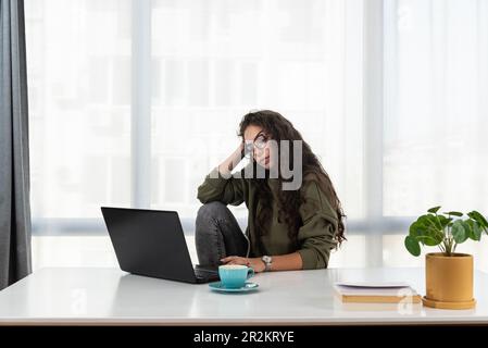 Gestresste junge Frau, die den Kopf in den Händen hält und sich demotiviert fühlt, während sie im Heimbüro sitzt und Remote mit einem Laptop arbeitet. Depressive Frauen Stockfoto