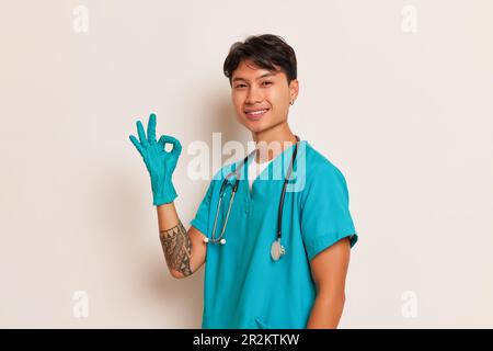 Junger Mediziner mit Stethoskop am Hals und Latexhandschuhen an seinen Händen posiert an der weißen Wand und zeigt mit einer Hand ein ok-Zeichen, professionell Stockfoto