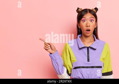 Ein überraschtes Mädchen mit zwei Brötchen in einem stylischen, sportlichen Sweatshirt steht auf pinkfarbenem Hintergrund, hebt die Hand hoch und zeigt rechts Stockfoto