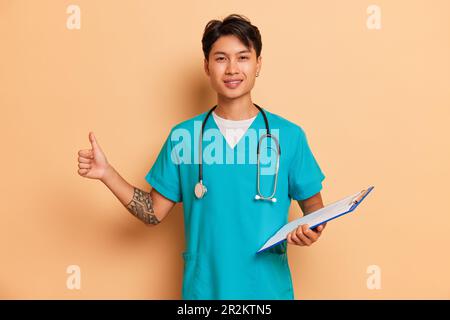 Lächelnder junger Mediziner mit Stethoskop am Hals posiert im Studio und hält das Klemmbrett in der Hand, professionelles Menschenkonzept, Kopierraum Stockfoto