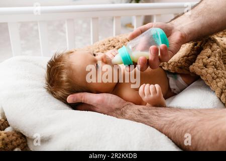 Vater behaarte Hände füttern Säugling mit Milchnahrung aus Plastikflasche, Neugeborenes bereitet sich auf Schlafenszeit vor Stockfoto