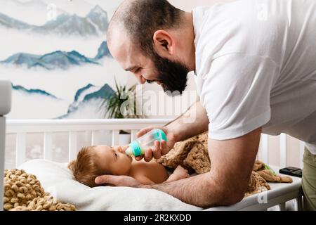 Der Vater füttert das Neugeborene mit Milch in einer Plastikflasche, Säuglingsnahrung, liegendes Kind im Kinderbett, Papa hält den Kopf fest, pflegt die Familie und liebt Stockfoto