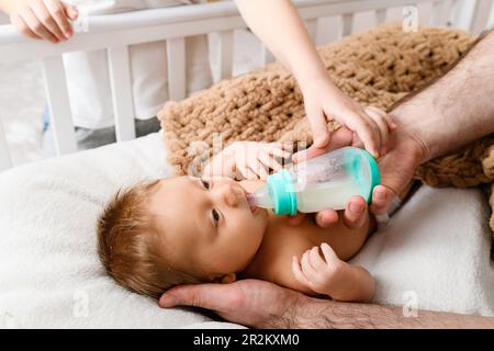 Ernährung von Säuglingen mit Milchnahrung in umweltfreundlichen Plastikflaschen, Hände des Vaters und älteres Kind halten Nahrung für schläfrige Neugeborene Stockfoto