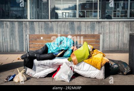 Armer müder, depressiver, hungriger Obdachloser oder Flüchtling, der auf einer Holzbank auf der Stadtstraße schläft, soziales Dokumentarkonzept Stockfoto