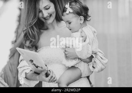 Schwarzweißfoto einer jungen blonden, eleganten Mutter, die ihr kleines Mädchen hält und ihr ein Buch zeigt Stockfoto