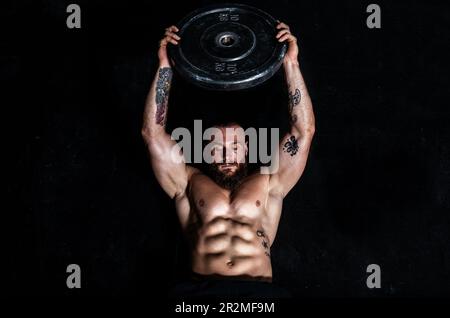 Junger aktiver, starker, schweißtreibender, fokussierter Mann mit großen Muskeln hält schwere Kugelhantel mit Heckengewichten für ein anstrengendes Cross-FIT-Training Stockfoto