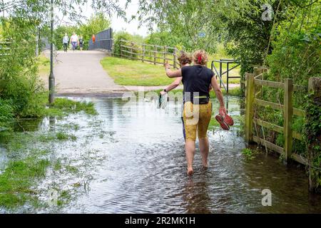 Eton Wick, Windsor, Großbritannien. 20. Mai 23. Jogger ziehen ihre Sportschuhe auf einem öffentlichen, überfluteten Fußweg aus. Der Roundmoor Graben Bach in Eton Wick ist wieder überflutet und bietet Wanderern Zugang zum Jubilee River Fußweg von Eton Wick, der nur mit dem Fahrrad oder in wellington Stiefeln zugänglich ist. Die nahegelegene Brücke über den Jubilee River wird regelmäßig von Einheimischen genutzt, die nach Asda in Cippenham gehen, um ihre Lebensmittel einzukaufen. Der Roundmoor-Graben ist bereits übergelaufen, als das Wasser der Themse angeblich überschüssiges Wasser aus den Kläranlagen der Themse in den Fluss gepumpt hat. Kredit: Maureen MCL Stockfoto