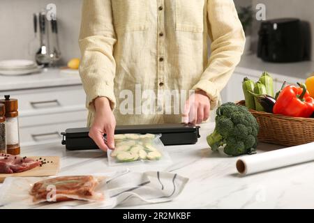 Frau, die eine Schließvorrichtung für Vakuumverpackungen benutzt, mit einer Plastiktüte Zucchini am weißen Tisch in der Küche, Nahaufnahme Stockfoto