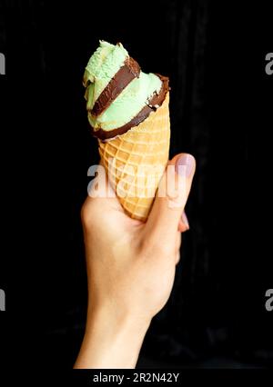 Die Hand hält weiches Eis in einem Waffelbecher mit Minze-Schokolade. Auf schwarzem Hintergrund, Nahaufnahme Stockfoto