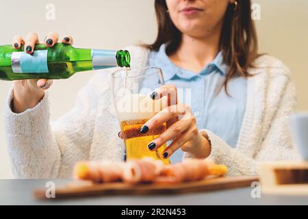 Unidentifizierbare Frau, die Bier aus der grünen Flasche in Glas gießt. Wurstplatte im Vordergrund verschwommen. Konzentrier dich auf Bierglas. Stockfoto