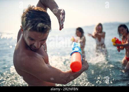 Drei Mädchen greifen einen Mann mit Wasserpistolen an. Eine Gruppe junger Freunde, die mit Wasserknaben im Wasser, im Meer spielen. Zusammensein, Spaß, Urlaub, Lifestyle CO Stockfoto