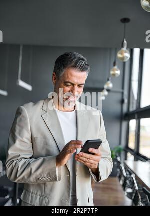 Glücklicher Geschäftsmann mittleren Alters, der im Büro ein Mobiltelefon in der Hand hält. Stockfoto