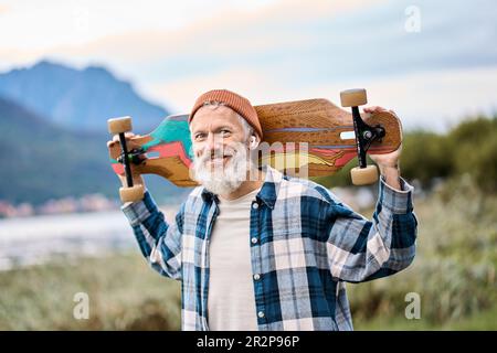 Ein älterer, glücklicher Hipster-Skater steht im Naturpark und hält Schlittschuh. Stockfoto