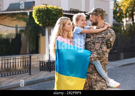 Soldat in Militäruniform wiedervereint mit seiner Familie und ukrainischer Flagge auf der Straße der Stadt, Platz für Text Stockfoto