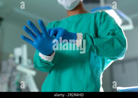 Mittelschnitt einer birassischen Chirurgin in Gesichtsmaske beim Anlegen von Operationshandschuhen im Operationssaal Stockfoto