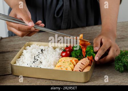 Kimchi mit Chopstick in koreanische Lunchbox mit Reis, Gyeranmari-Eierrolle, Tomaten, Brokkoli und schwarzem Sesamsamen geben. Stockfoto