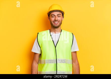 Fröhlicher, bärtiger Arbeiter mit gelbem Schutzhelm und gelber Weste steht mitten auf gelbem Hintergrund und lächelt, professionelles Menschenkonzept, Text Stockfoto