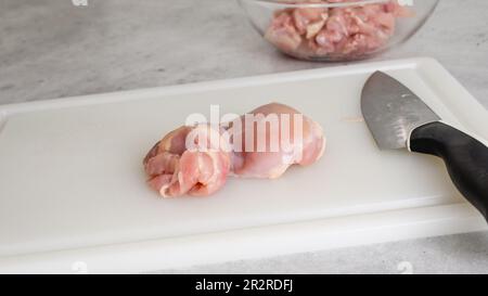 Hähnchenschenkel und Küchenmesser Nahaufnahme auf einem weißen Kunststoffschneidbrett auf einem Küchentisch. Mariniertes Hühnerfilet, Nahzubereitung p Stockfoto