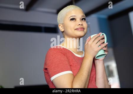 Fröhliche, modische, unveränderte, birassische Frau, die mit Kaffee steht und zu Hause im Wohnzimmer lächelt. Entspannung, Freizeit und häuslicher Lebensstil. Stockfoto