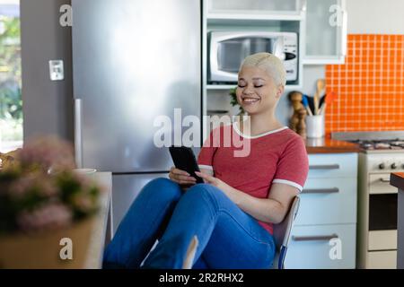 Glückliche birassische Frau, die am Tisch sitzt und ihr Smartphone in der Küche benutzt. Lebensstil, Kommunikation und häusliches Leben. Stockfoto