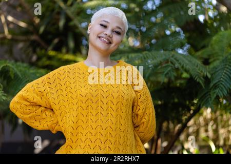 Unverändertes Porträt einer modischen, birassischen Frau im gelben Pullover, die im Garten lächelnd steht. Entspannung, Freizeit, Natur und Lebensstil. Stockfoto