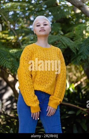 Unverändertes Porträt einer fröhlichen, modischen, birassischen Frau im gelben Pullover, die im Garten an Bäumen steht. Entspannung, Freizeit, Natur und Lebensstil. Stockfoto