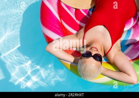Lächelnde, unveränderte, birassische Frau in Sonnenbrille beim Sonnenbaden auf einem aufblasbaren Schwimmbad. Sommer, Freizeit, Entspannung und Urlaub. Stockfoto