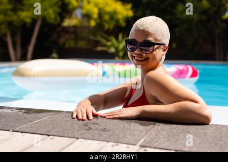 Porträt einer glücklichen, unveränderten, birassischen Frau mit Sonnenbrille, die am Pool steht. Sommer, Freizeit, Entspannung und Urlaub. Stockfoto