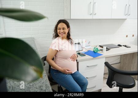 Porträt einer glücklichen schwangeren Frau, die lächelt und ihren Bauch hält, Besuch einer medizinischen Klinik zur Schwangerschaftskontrolle Stockfoto