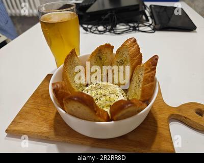 Im Ofen gebackener französischer Camembert-Käse mit Rosmarin-Zweigen, Knoblauchzehen und Lorbeerblättern, serviert mit Croutons auf einem Holzbrett auf einem dunklen Holzbao Stockfoto