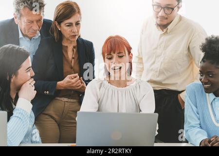 Eine junge Frau sitzt an ihrem Schreibtisch vor einem Laptop und präsentiert ein Projekt. Ihre Kollegen, eine multigenerationale und multirassische Gruppe, passen auf Wit auf Stockfoto