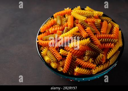 Trockene Nudeln mit Fusilli auf dunklem Hintergrund. Dreifarbige Fusilli-Pasta in einer Schüssel. Draufsicht. Speicherplatz kopieren Stockfoto