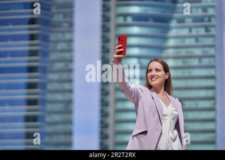 Eine lächelnde Geschäftsfrau, die im Finanzdistrikt draußen mit einem Mobiltelefon Selfies macht. Technologie und Geschäftskonzept. Stockfoto