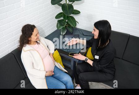 Draufsicht der schwangeren Frau, die ihren Bauch berührt, auf dem Sofa in der medizinischen Klinik sitzt, Beratung mit dem Geburtshelfer Stockfoto