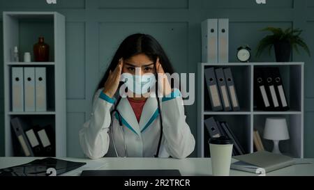 Ungesunde müde kranke Ärztin junge Frau in der medizinischen Maske berühren Kopfbügel leiden an Kopfschmerzen müde enttäuscht weibliche medizinische Krankenschwester fühlen Stress Stockfoto