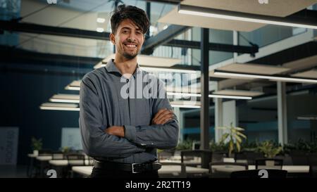 Ein erfolgreicher lächelnder indisch-arabischer Geschäftsmann, der im Amt mit gekreuzten Armen posierte. Selbstbewusster, männlicher Agent, Firmeninhaber, Geschäftsführer, jung Stockfoto