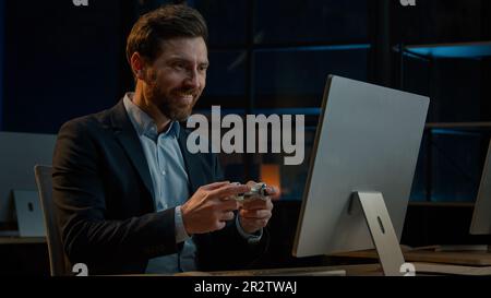 Ein erwachsener Geschäftsmann, der im Abendbüro arbeitet, glücklicher männlicher Gamer, ein weißer Mann, macht Pause und spielt Konsolenvideospiel auf einem Computer mit Spielen Stockfoto