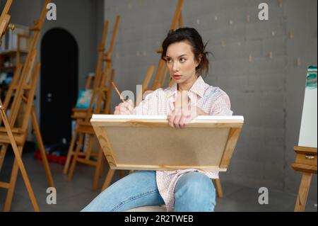 Attraktive junge Künstlerin, die Leinwand in der Hand hält und ein Bild zeichnet Stockfoto