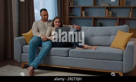 Ein vielseitiges, multiethnisches Paar, das auf dem Sofa sitzt und mit dem Computer zu Hause auf der Website surft, entspannende Frau und Mann, die sich im Einzelhandel unterhalten Stockfoto