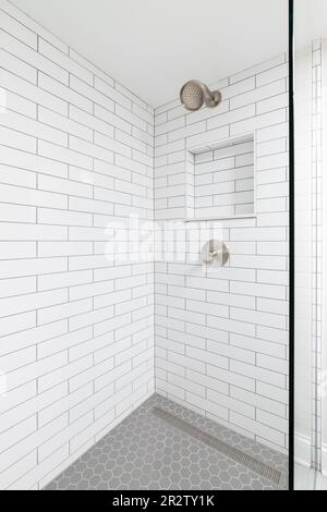 Eine Badezimmerdusche mit weißen Fliesenwänden aus der U-Bahn, ein eingebautes Nischenregal, ein Bronze-Duschkopf und ein grauer sechseckiger Fliesenboden. Stockfoto